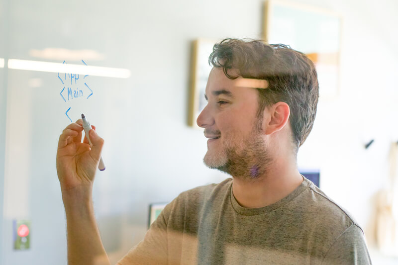 A smiling man writes a sample app structure on a glass wall with a dry erase marker.