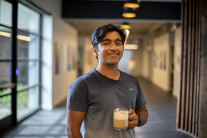 A smiling man holding a mug of foamy latte is in a warmly lit hallway